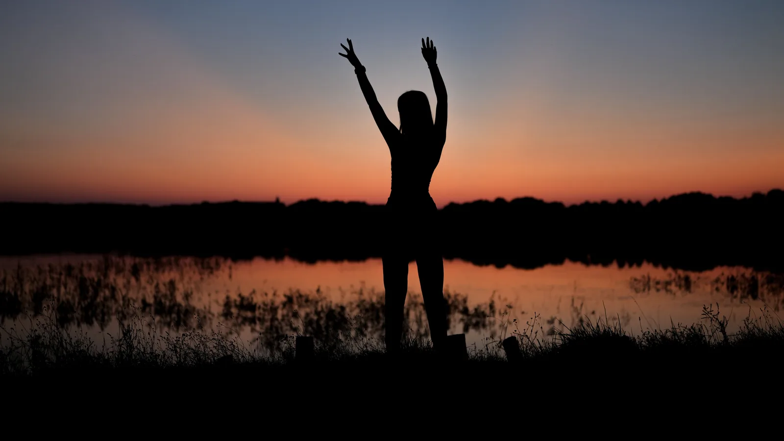 Silhouet van een vrouw met geheven armen aan de oever van een meer bij zonsondergang