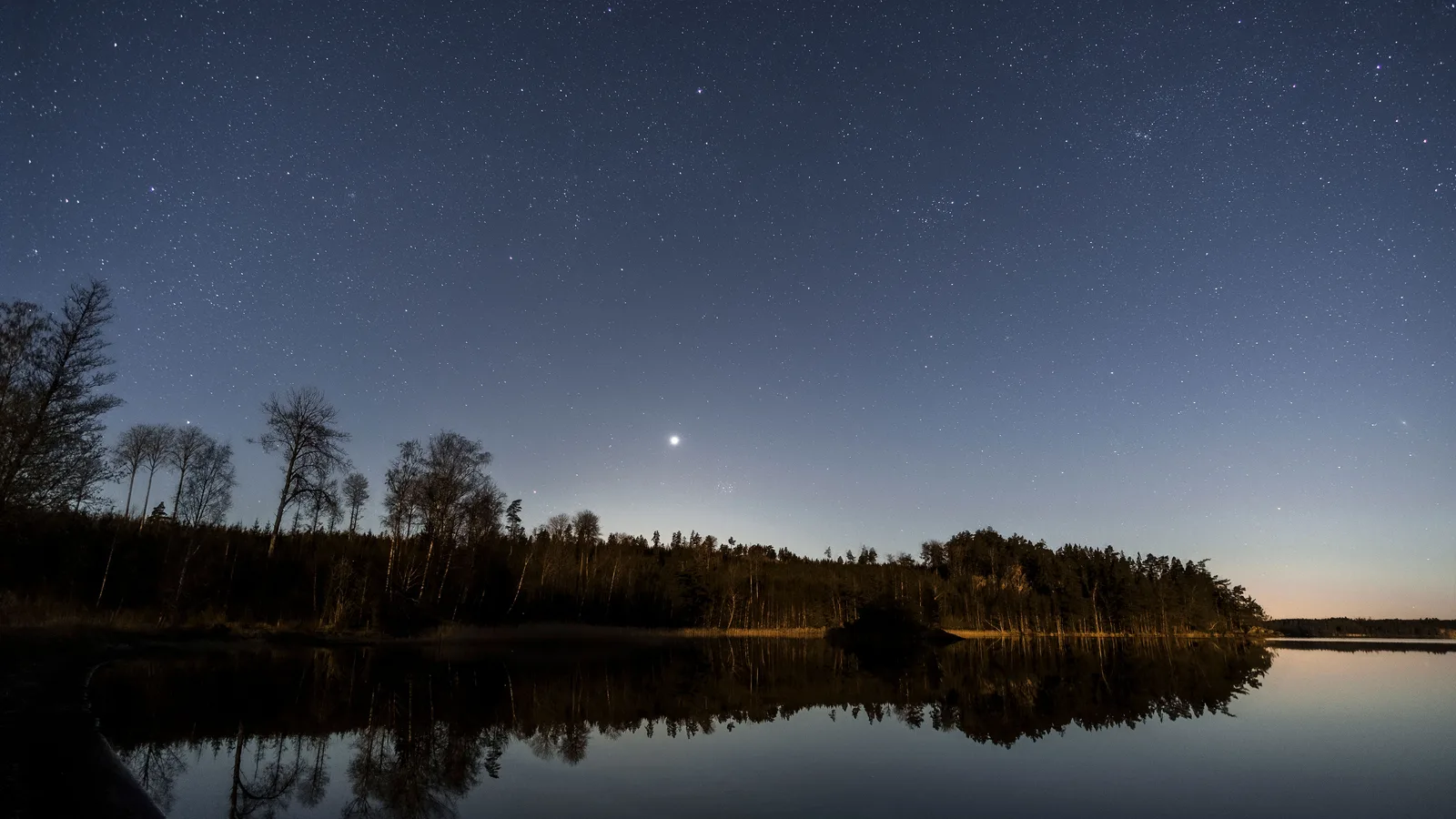 Een heldere planeet staat boven een donkere bosrand en weerspiegelt in het stille meer
