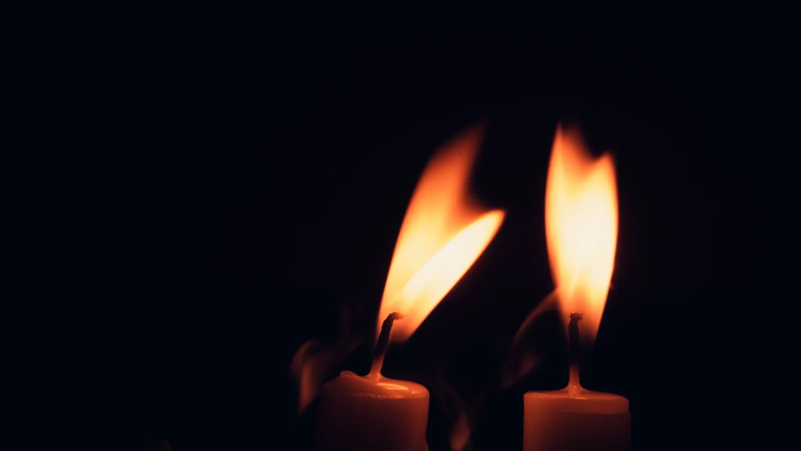 Twee brandende kaarsen naast elkaar in het donker, de vlammen buigen naar elkaar toe
